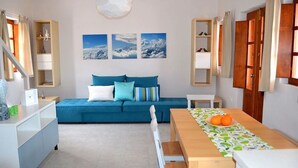 Una sala moderna con un sofá azul, una mesa de comedor de madera con un centro de mesa de frutas y una zona de cocina con armarios y una lámpara colgante.