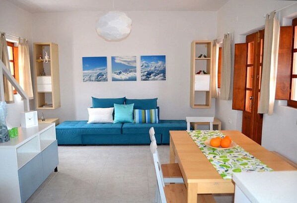 A modern living room with a blue sofa, a wooden dining table with a fruit centerpiece, and a kitchen area with cabinets and a hanging light fixture. - Cactus hideaway with sea views (Imerovigli)