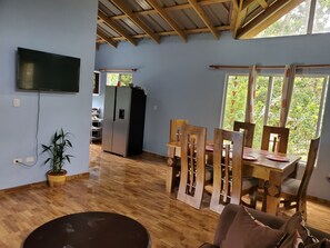 A dining area with wooden chairs and a table, a kitchen with a refrigerator, and a TV mounted on the wall.