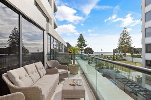 A balcony with wicker furniture, a glass railing, and a view of the sea and trees. - Marvellous Mount Apartment with pool, gym & beach (Mount Maunganui)