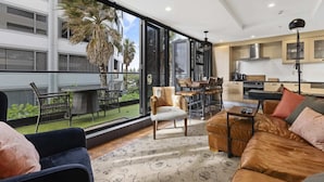 A modern living room with a large window, a sofa, a coffee table, and a dining area with chairs. - Marvellous Mount Apartment with pool, gym & beach (Mount Maunganui)