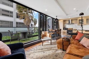 A modern living room with a large window, a sofa, a coffee table, and a dining area with chairs. - Marvellous Mount Apartment with pool, gym & beach (Mount Maunganui)