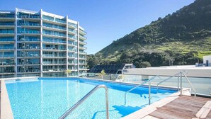 A swimming pool with a wooden deck, a modern hotel building, and a hilly landscape in the background. - Marvellous Mount Apartment with pool, gym & beach (Mount Maunganui)