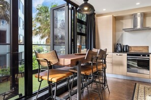A modern kitchen with a dining area, large windows, and a view of greenery. - Marvellous Mount Apartment with pool, gym & beach (Mount Maunganui)