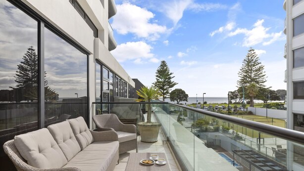 A balcony with wicker furniture, a glass railing, and a view of the sea and trees. - Marvellous Mount Apartment with pool, gym & beach (Mount Maunganui)