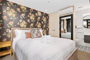 A neatly made bed with white linens and decorative pillows, a wooden nightstand, and a floral wallpaper. - Marvellous Mount Apartment with pool, gym & beach (Mount Maunganui)