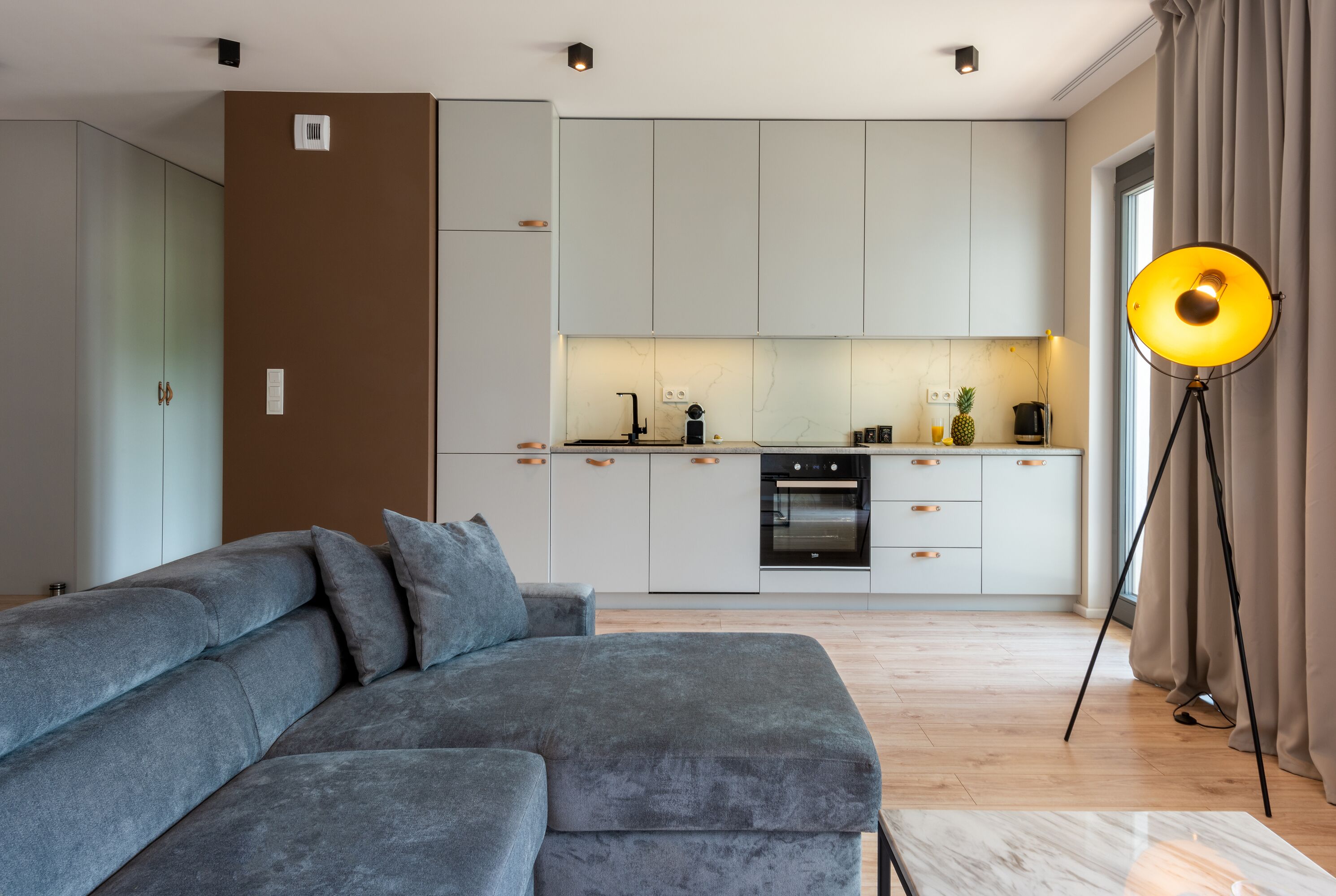 Un salon moderne avec un canapé gris, une cuisine avec des meubles blancs et un lampadaire.