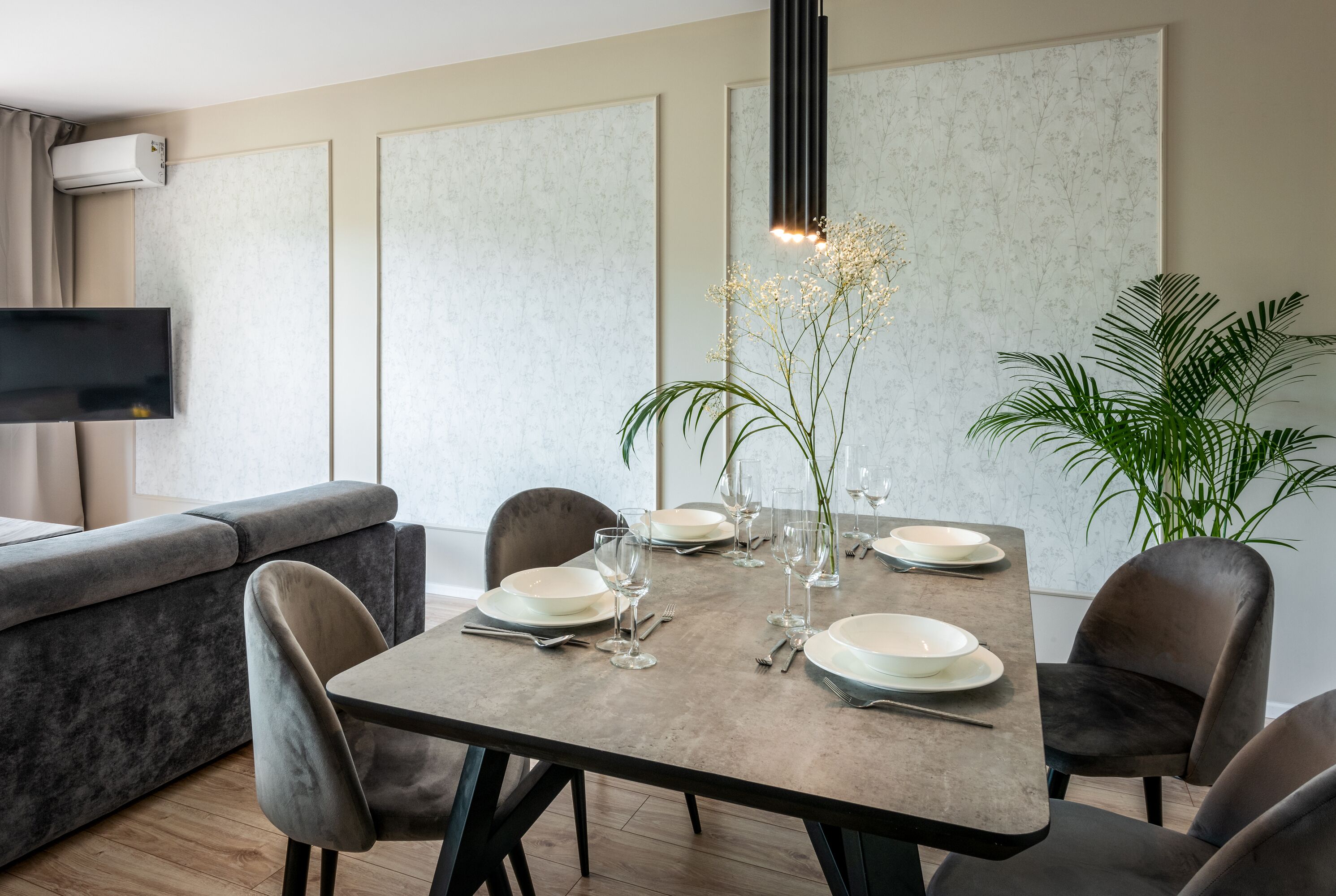 Een moderne eetruimte met een rechthoekige tafel voor zes personen, omringd door zes stoelen. Rechts staat een grote potplant en links is een flatscreen televisie aan de muur bevestigd.
