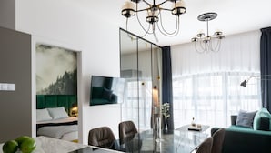 Habitación de hotel moderna con una mesa de comedor de cristal, asientos cómodos, un sofá verde, un espejo grande y un cuadro de un bosque.