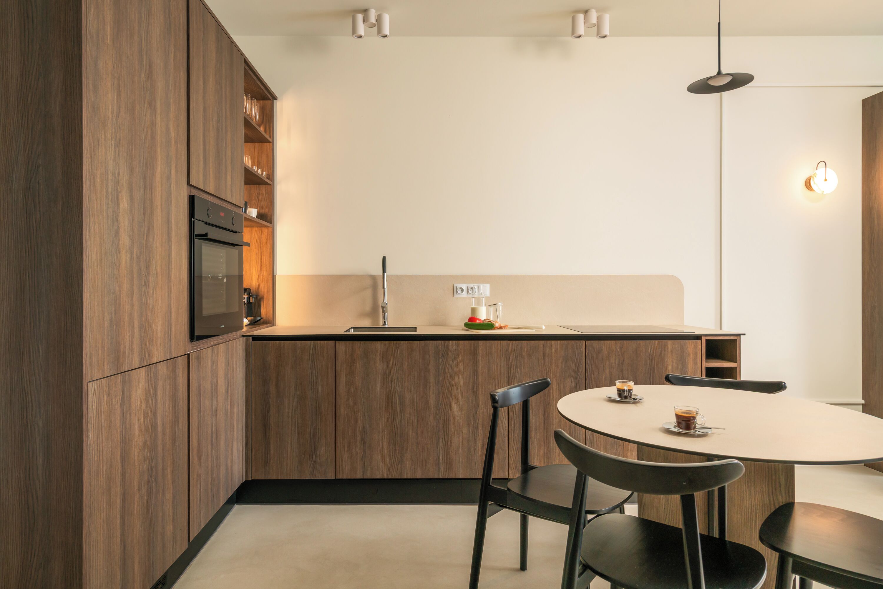 Une cuisine moderne avec des meubles en bois, un four encastré, une table à manger avec des chaises et des luminaires suspendus.