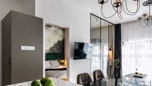 Una habitación de hotel moderna con una mesa de comedor de vidrio, una cama con cabecera verde, un televisor y un amplio ventanal con cortinas.