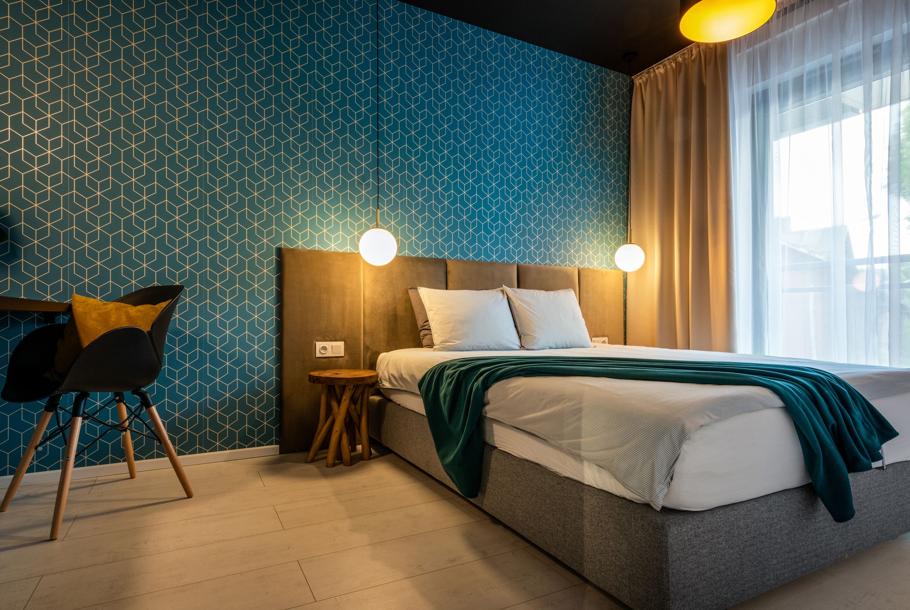 A modern hotel room with a geometric patterned wall, a large bed with white bedding and a green throw, a wooden chair, and a small side table.