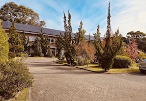 Um edifício residencial com entrada pavimentada, cercado por árvores e arbustos diversos.