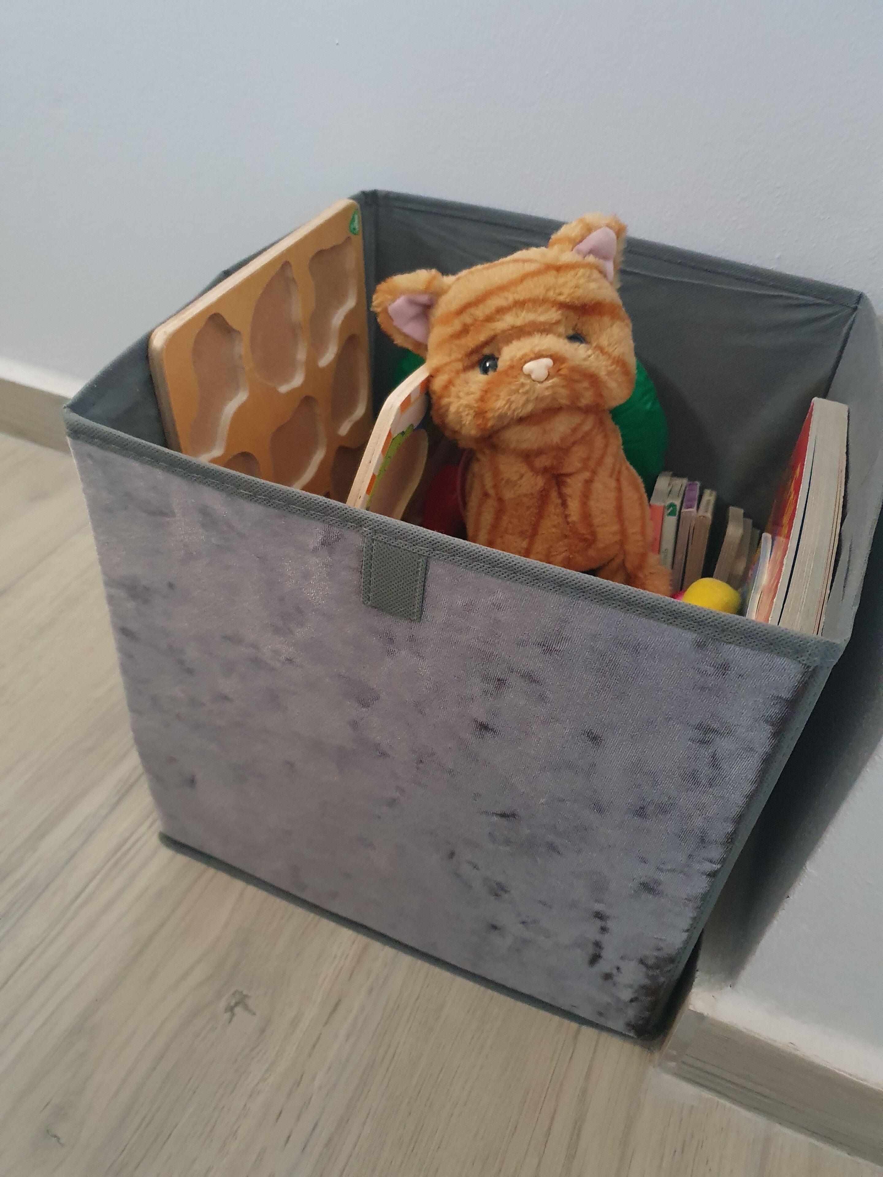 Harmaa säilytyslaatikko, jossa on kahva, ja sen sisällä on pehmeä pehmolelu ja joitain kirjoja.