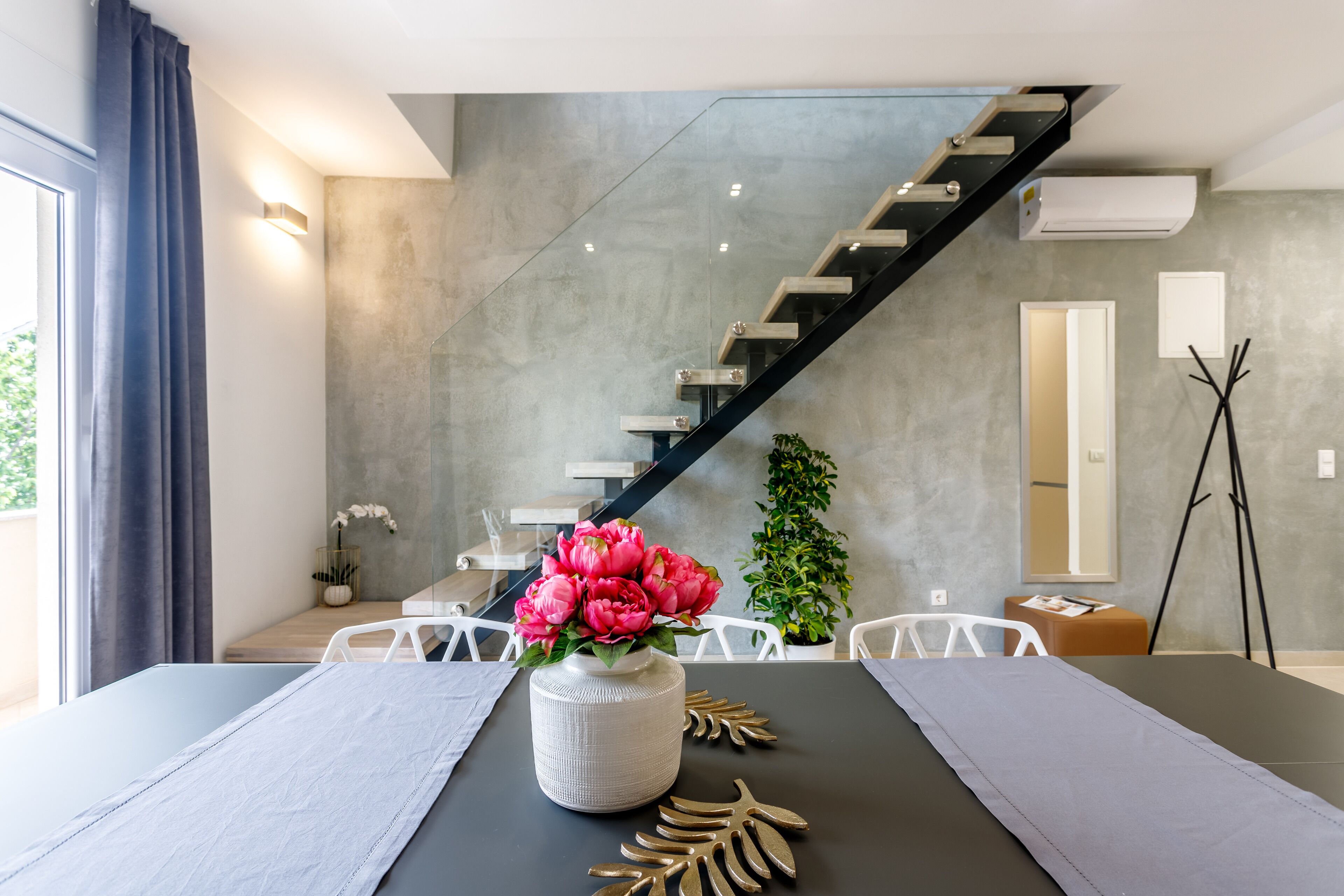 A modern dining area with a table set for two, a vase of flowers, and a staircase in the background.