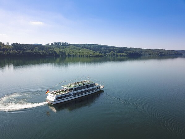 A river cruise ship with multiple decks and a flag, moving on calm water with a forested shoreline in the background. - House (Sundern (Sauerland))