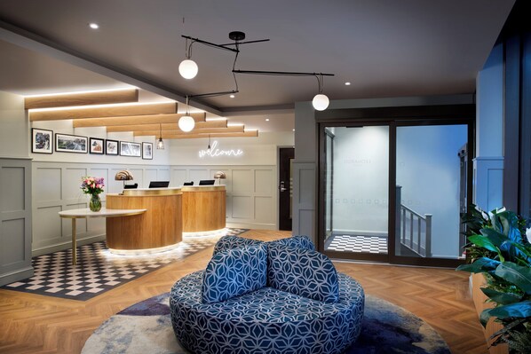 A modern reception area with a wooden reception desk, a 'welcome' sign, a seating area with patterned cushions, and a checkered floor. - Leonardo Hotel Chester (Chester)