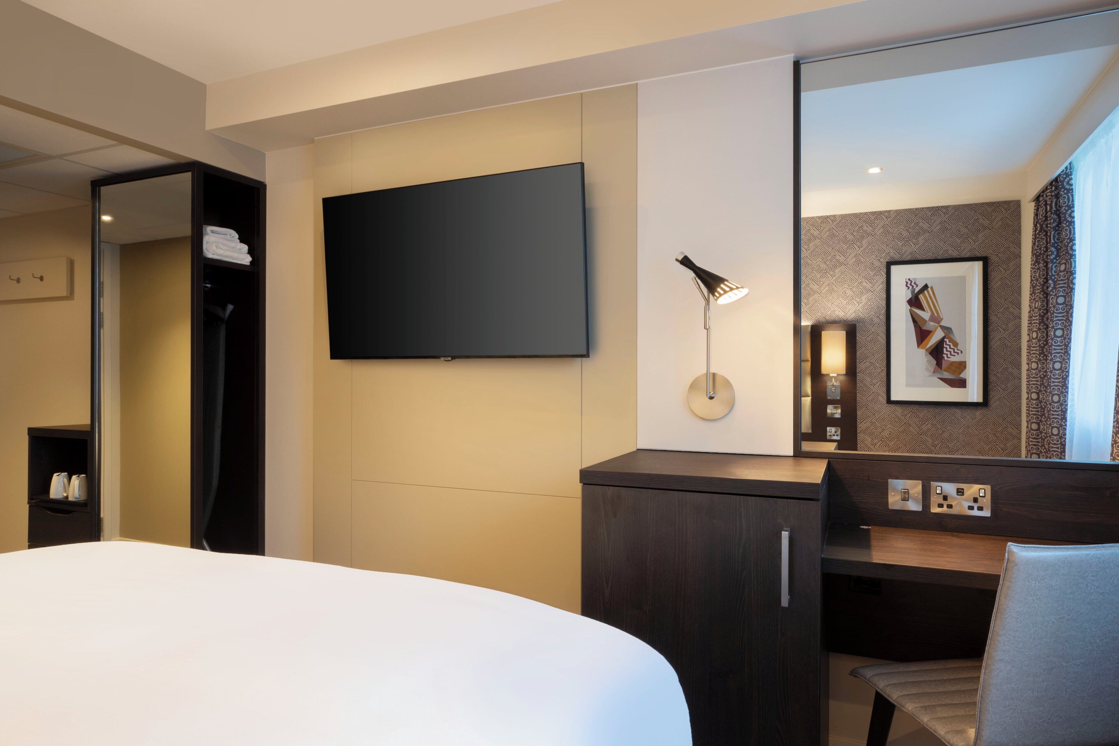 Quarto de hotel com cama, escrivaninha, televisão de tela plana na parede, espelho e uma obra de arte emoldurada.