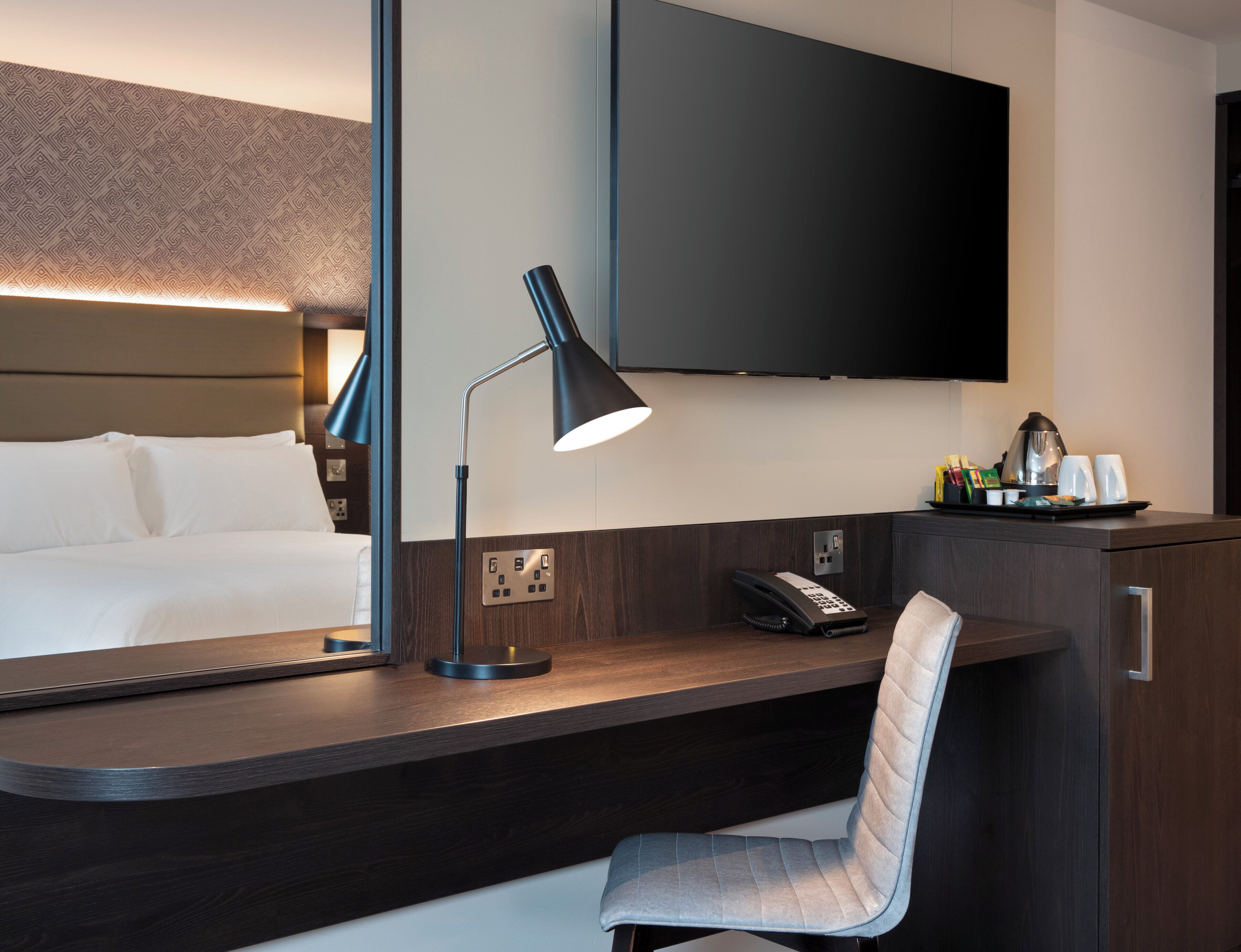 Ein modernes Hotelzimmer mit einem großen Bett, einem Schreibtisch mit Lampe, einem Sessel, einem an der Wand befestigten Flachbildfernseher und einem Telefon auf dem Schreibtisch.