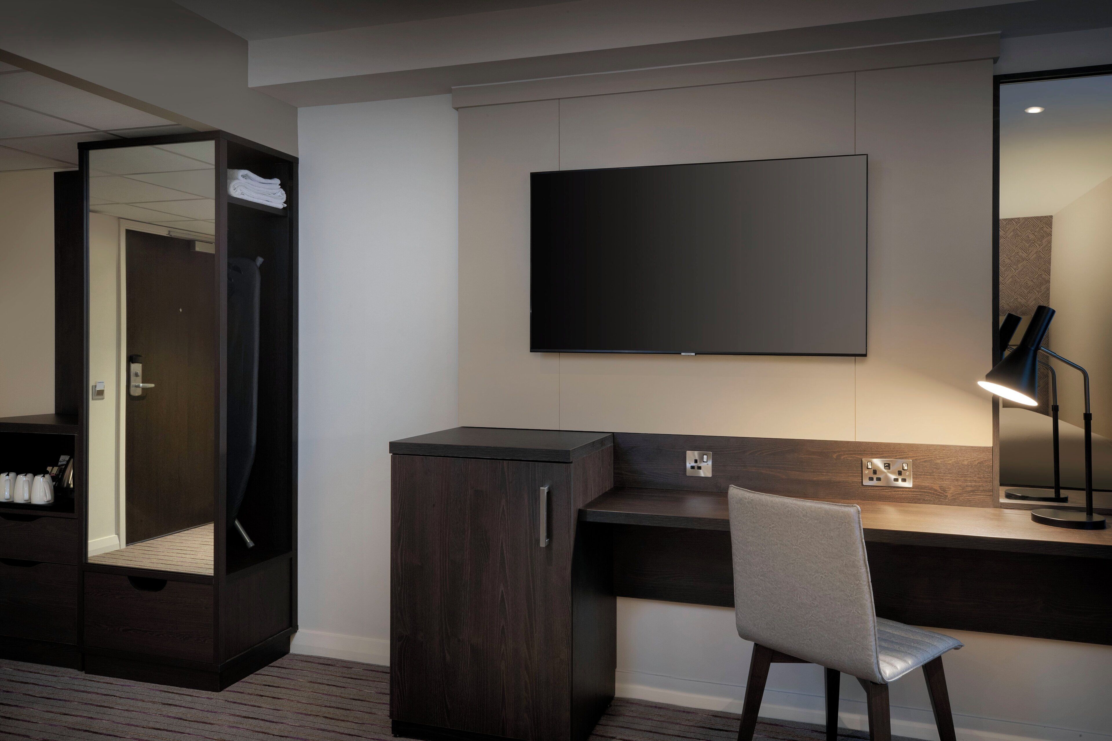 A modern hotel room with a dark wood wardrobe, a desk with a chair, a flat-screen TV, and a mirror.