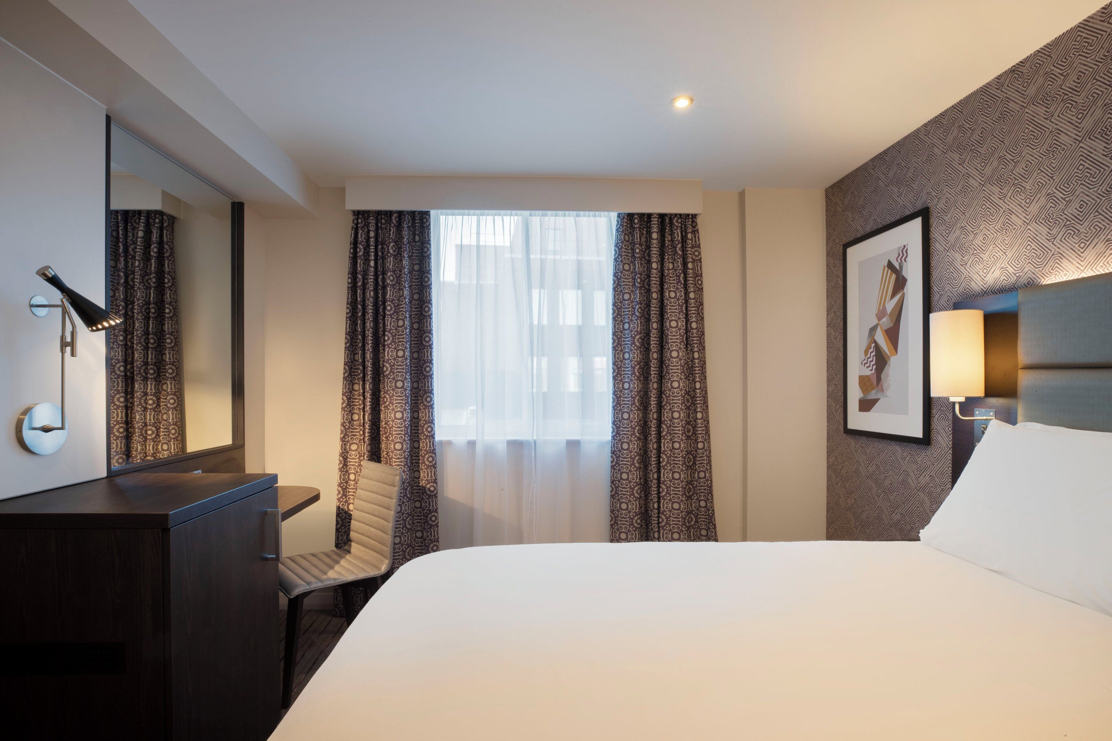 A hotel room with a large bed, a desk with a chair, a mirror, and a window with patterned curtains.