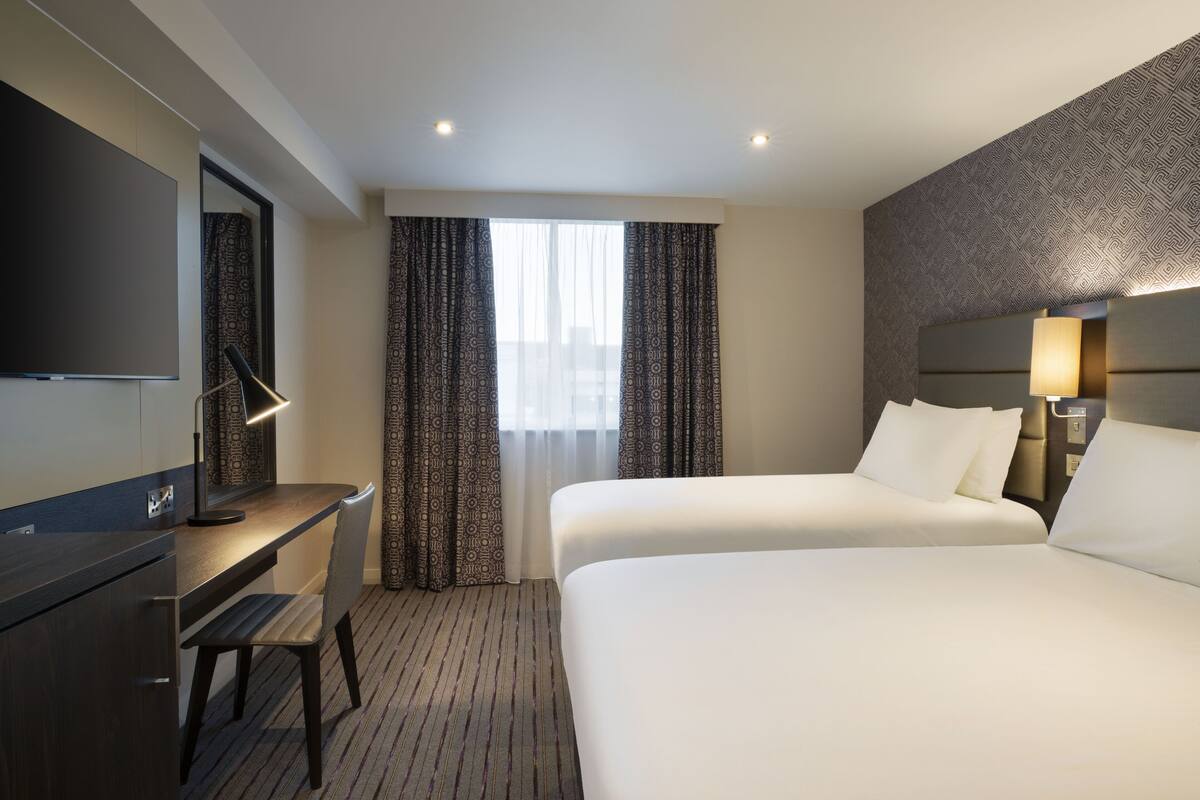 A hotel room with a bed, a desk, a chair, a TV, and a window with curtains.
