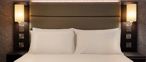 A neatly made bed with white linens, a headboard with a light fixture, and a patterned wallpaper.