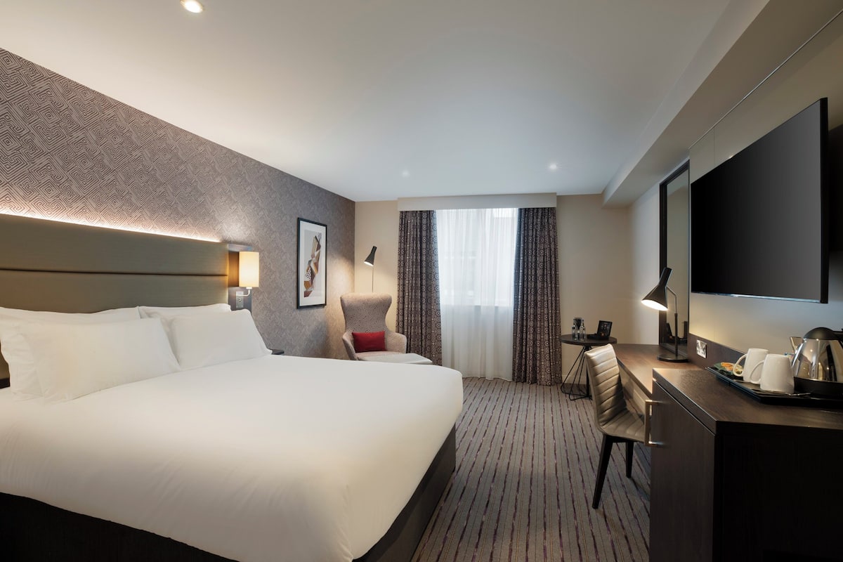 A hotel room with a large bed, a desk with a lamp, a chair, a TV, and a window with curtains.