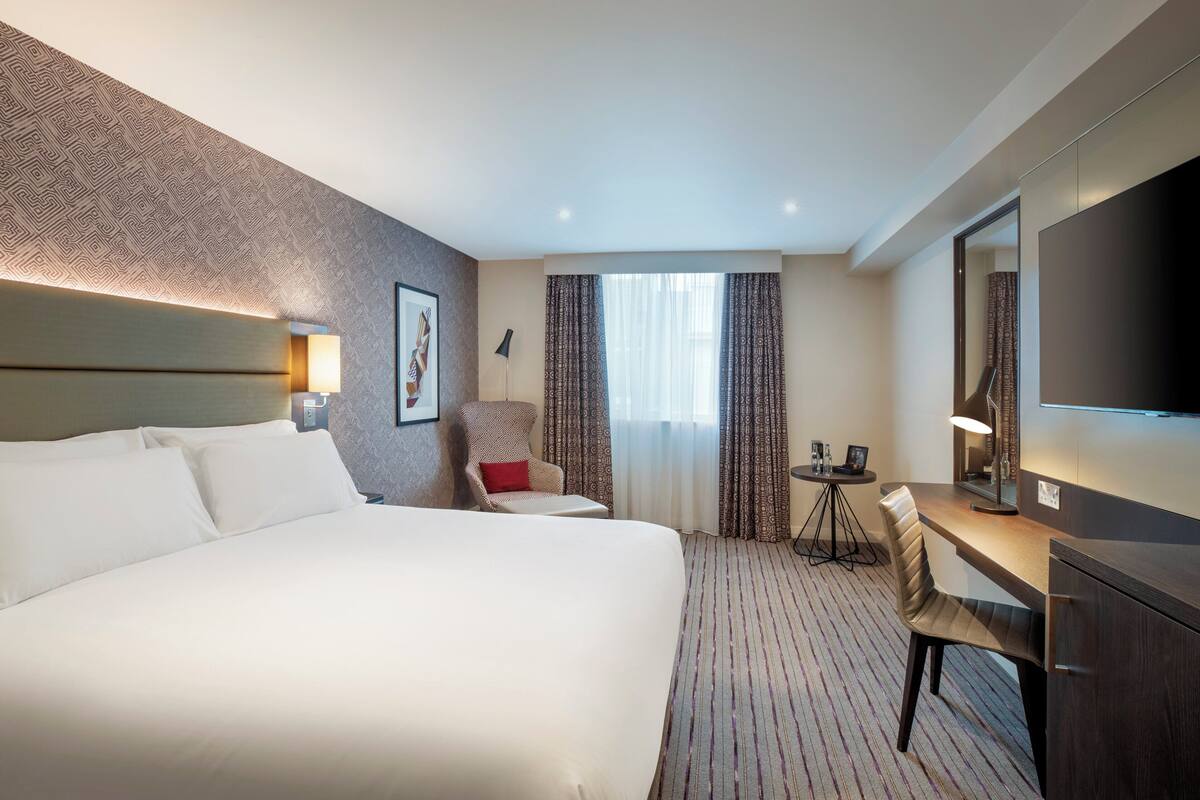 A hotel room with a large bed, a desk, a chair, a TV, and a small table with a lamp.