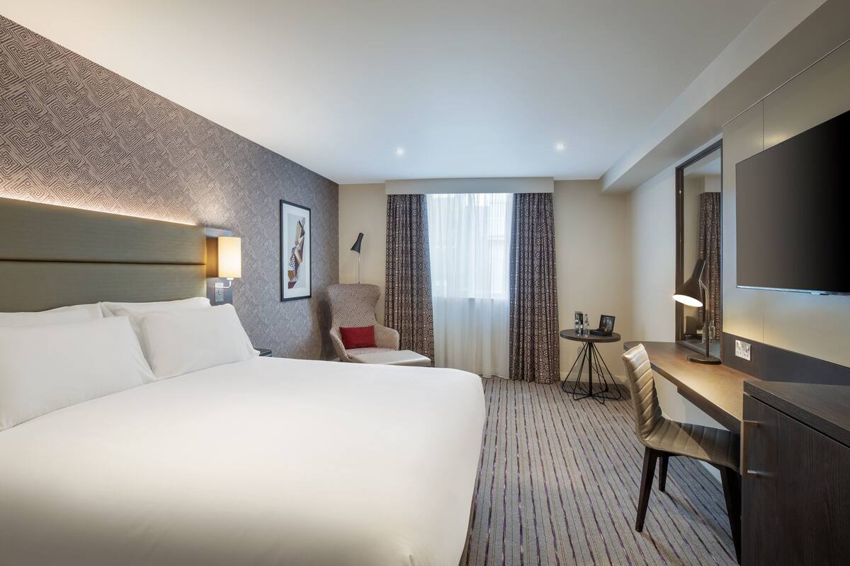 A hotel room with a large bed, a desk, a chair, a TV, and a small table with a lamp.