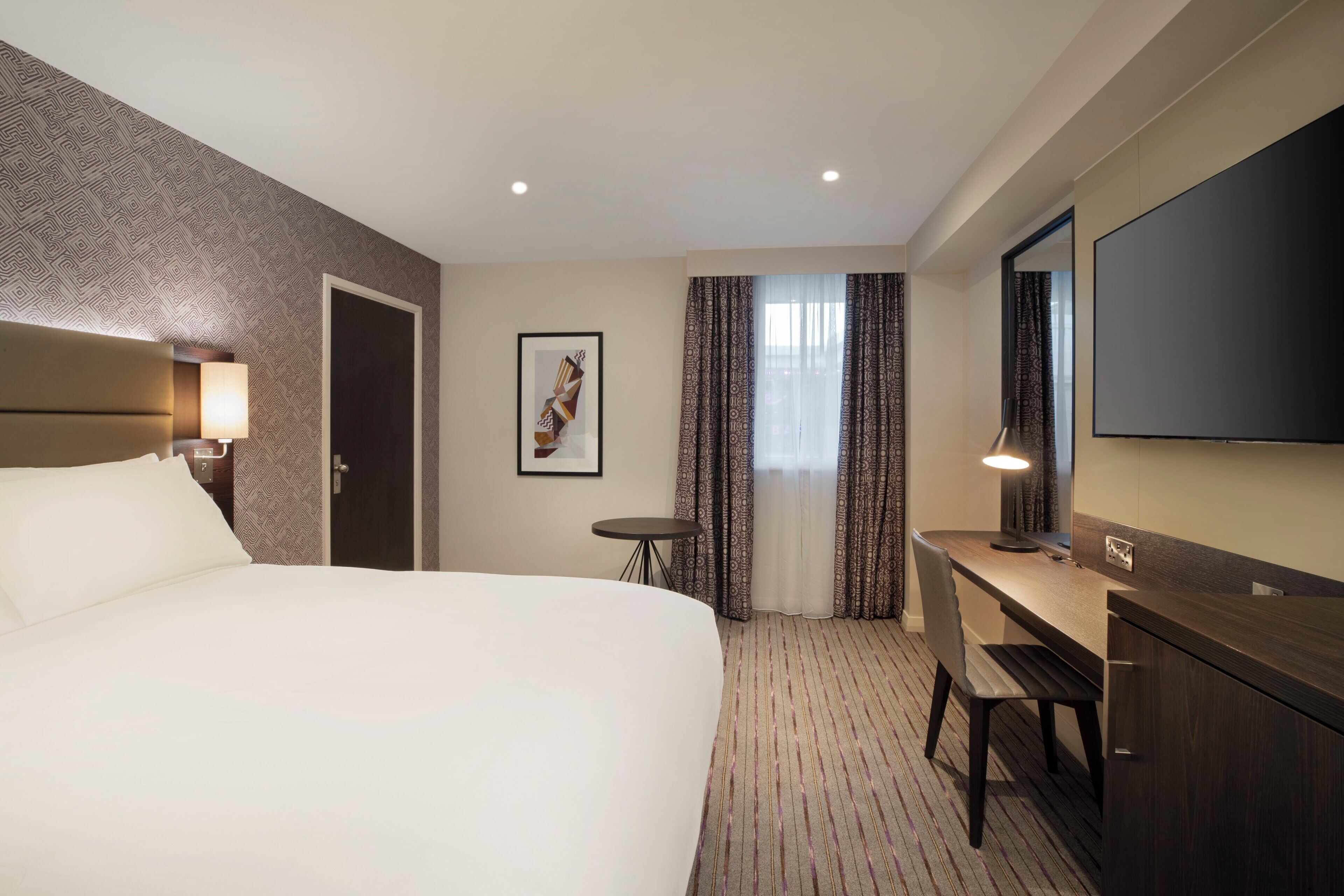 Hotelkamer met een groot bed, een bureau, een stoel, een kleine tafel, een lamp, een televisie en een behang met een patroon.