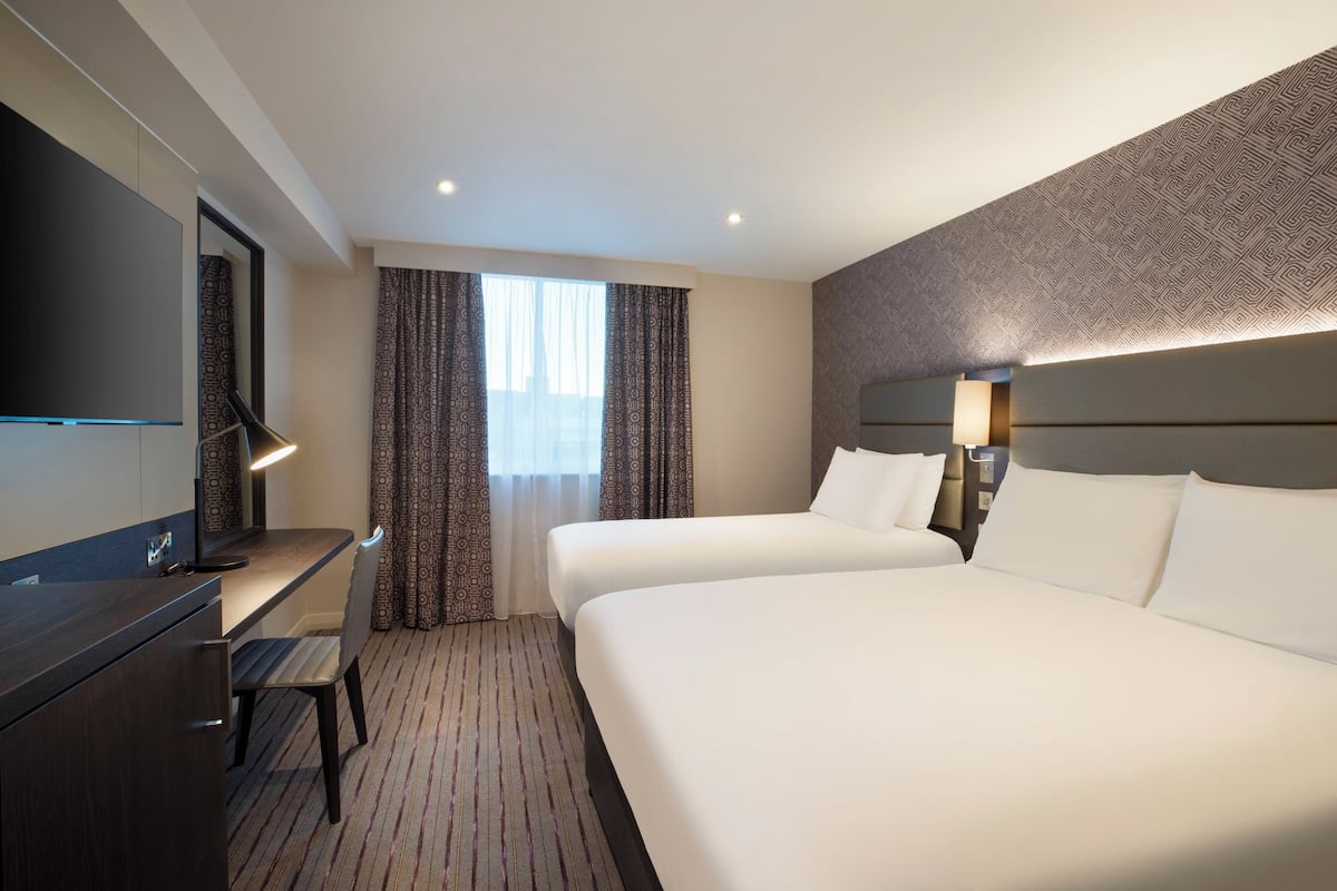 A hotel room with two beds, a desk, a chair, a TV, and a patterned wall.
