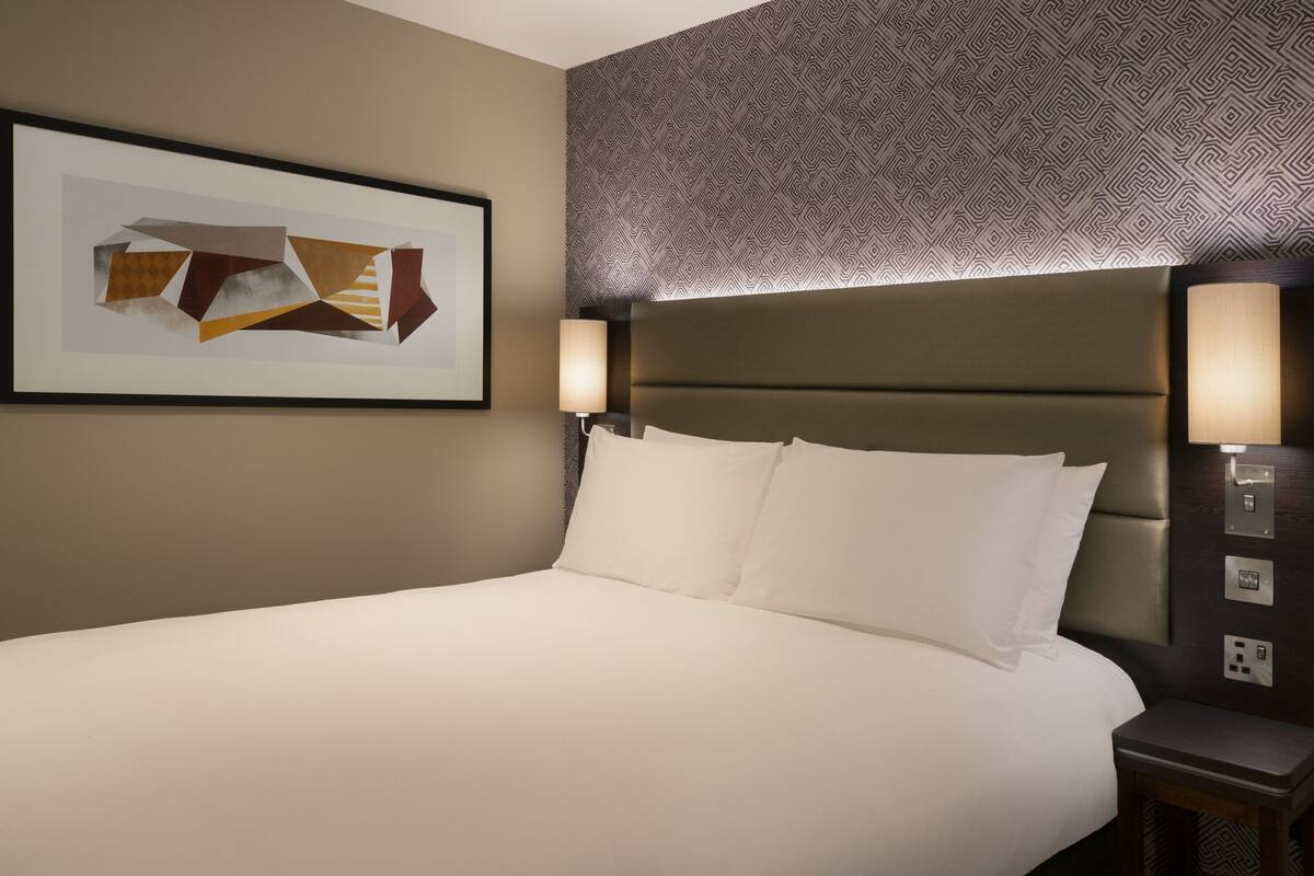 A hotel room with a large bed, two bedside lamps, a framed artwork on the wall, and a patterned wallpaper.