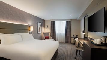 A hotel room with a large bed, a desk with a lamp, a chair, a TV, and a window with curtains.