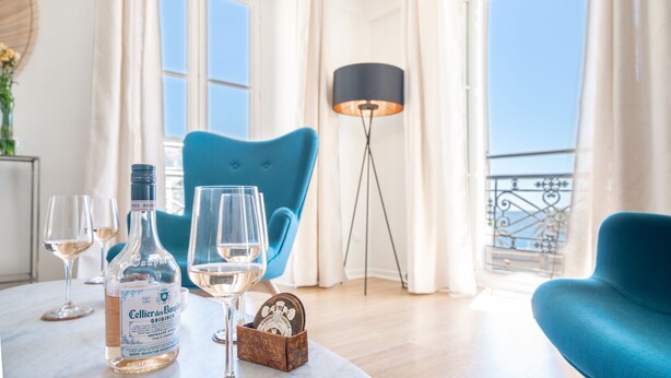 Une pièce avec une table mise, comprenant des verres à vin et une bouteille de Cellerie de Dauron. On y trouve également une chaise bleue et un lampadaire.