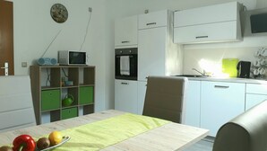 A compact kitchen with white cabinets, a microwave, a small oven, and a sink. There is a green shelf with kitchen items, a clock on the wall, and a chair in the dining area. - Large 90 sqm vacation apartment (Schönstedt)