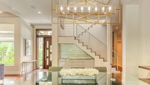 Un interior moderno con una mesa de centro de vidrio, un arreglo floral blanco, una escalera con barandilla de cristal y un candelabro geométrico.