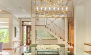 A modern interior with a glass coffee table, a white floral arrangement, a staircase with a glass railing, and a geometric chandelier.