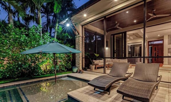 A pool area with lounge chairs and an umbrella, surrounded by lush greenery.