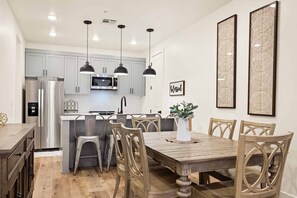 A modern kitchen with light grey cabinets, a stainless steel refrigerator, a microwave, and pendant lights. There is a dining area with a wooden table and chairs, and a potted plant on the table.