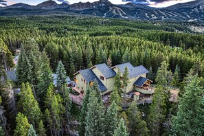 Une maison à deux étages, entourée d’une forêt dense, avec une chaîne de montagnes en arrière-plan.