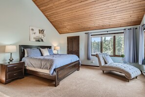 Ein Schlafzimmer mit Holzbalkendecke, einem Bett mit Kopfteil, einem Nachttisch mit Lampe, einer Liege und einem Fenster mit Vorhängen.