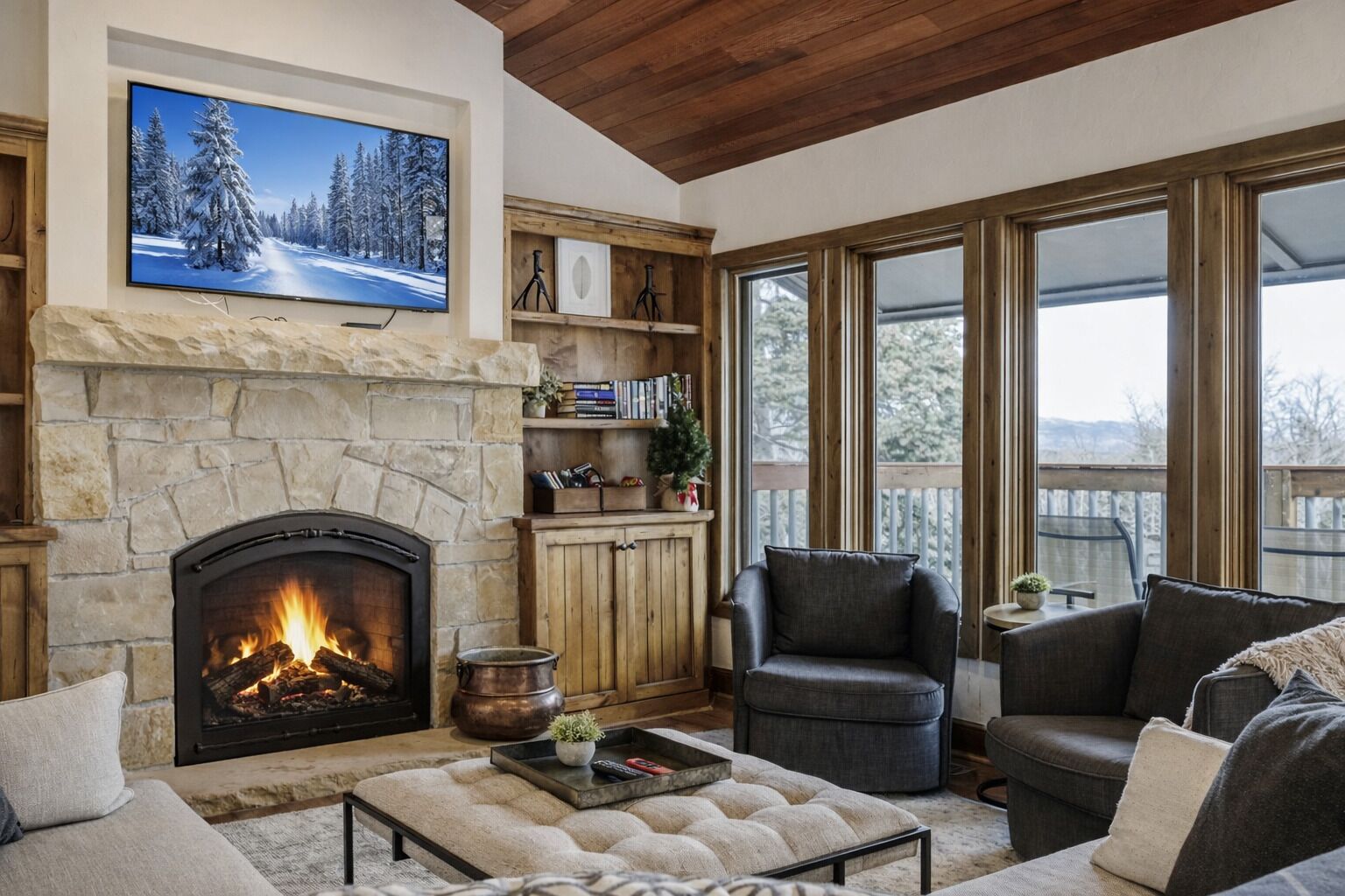 Una sala de estar acogedora con chimenea de piedra, un televisor montado encima y un ventanal con vistas a árboles.