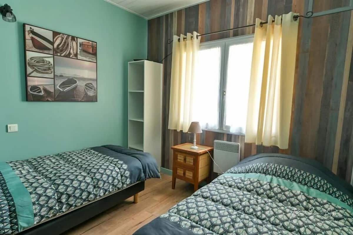 A room with two beds, a window with curtains, a wall-mounted shelf, and a wall art depicting boats.
