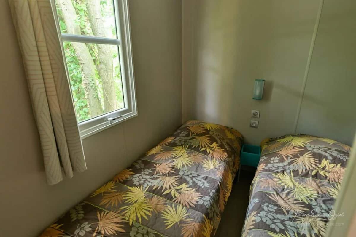 A small, single-room accommodation with a window providing a view of greenery outside. There are two single beds with floral-patterned bedding.