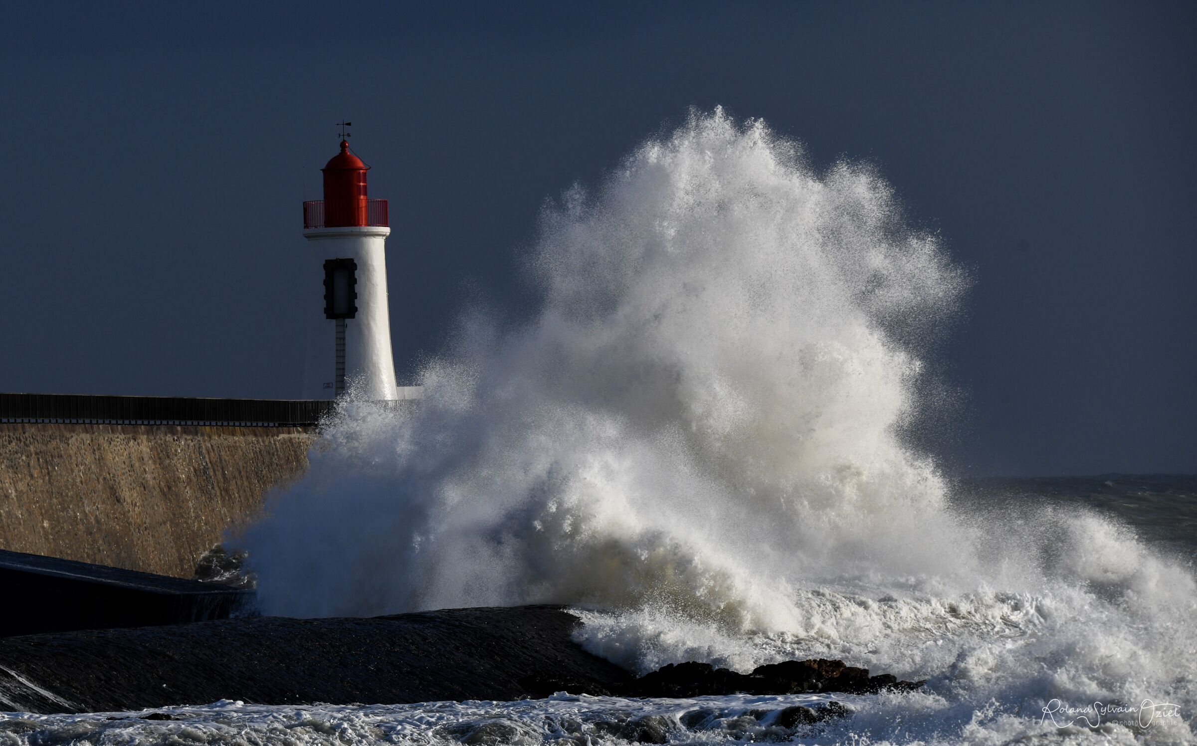 A lighthouse by a rocky shore with waves crashing against it.