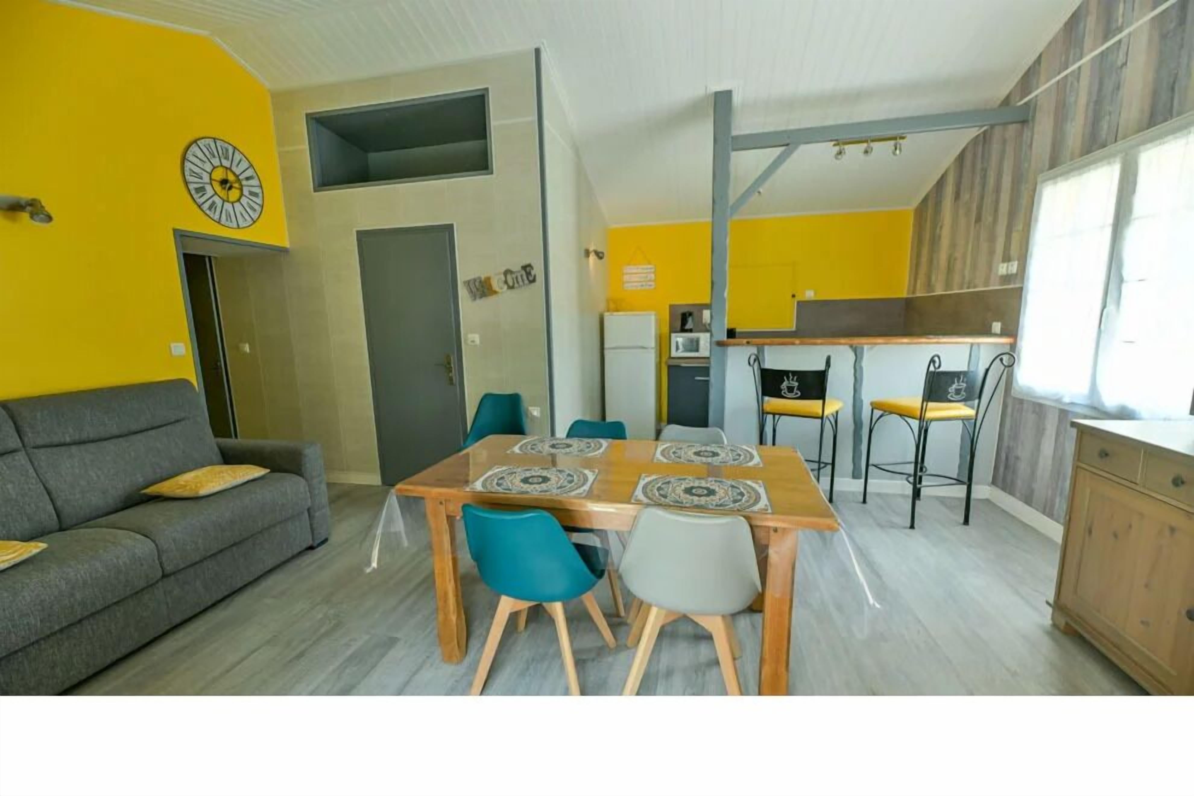 A modern living room with a sofa, a dining table with chairs, a kitchen area with a refrigerator and microwave, and a yellow accent wall.