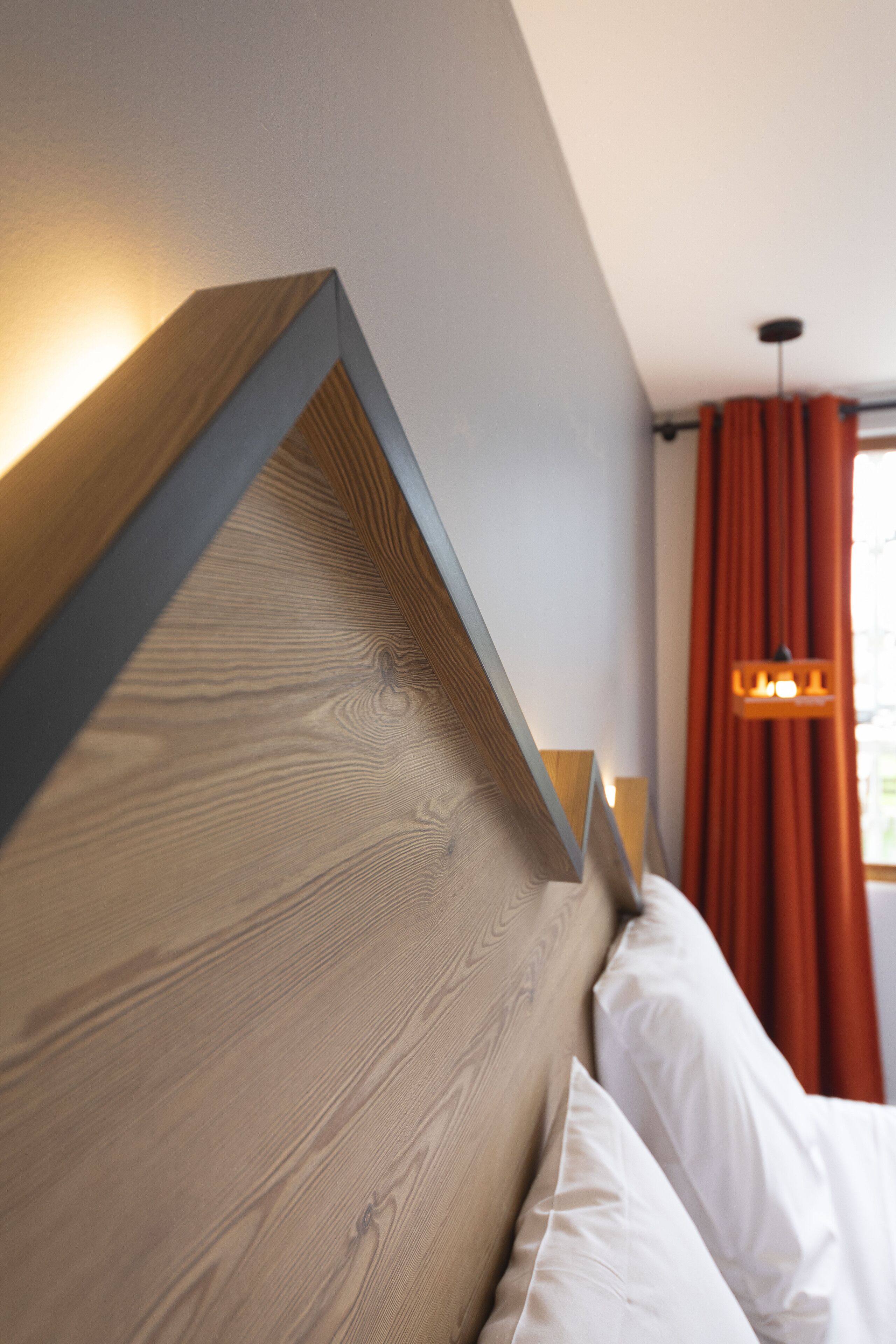 A modern bedroom with a wooden headboard, white bedding, and a window with orange curtains.
