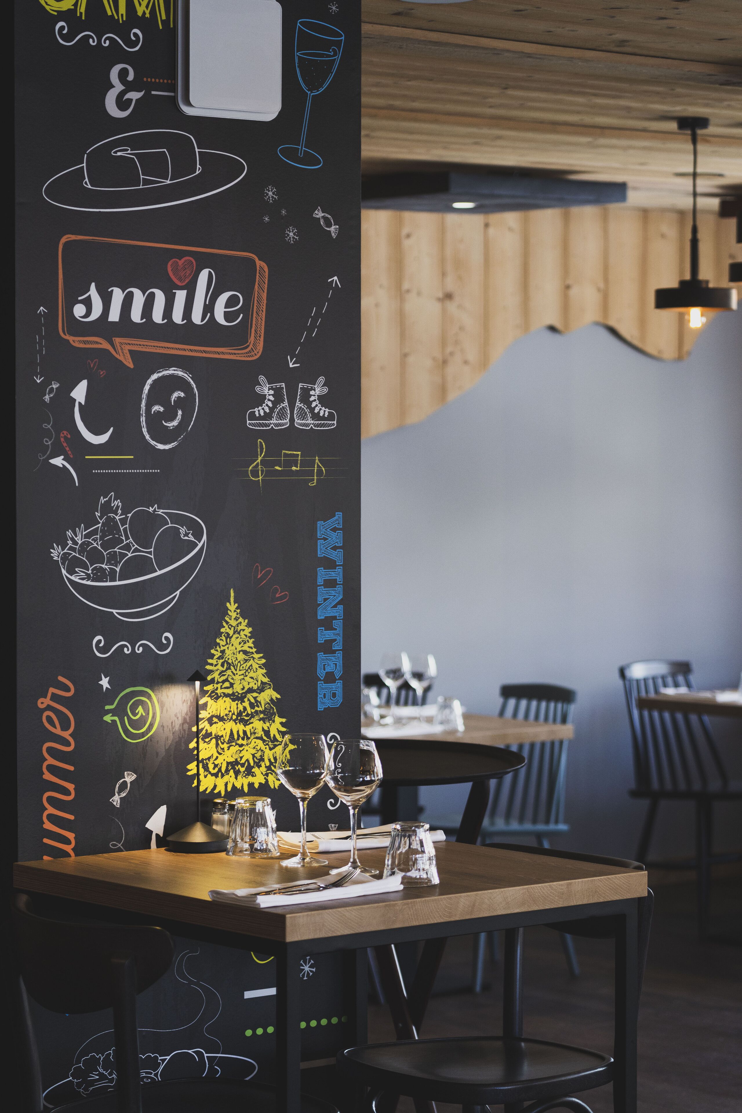En restaurantindretning med en tavlevæg med forskellige tegninger og ordet 'smile'. Bordene er dækket med vinglas og menukort.
