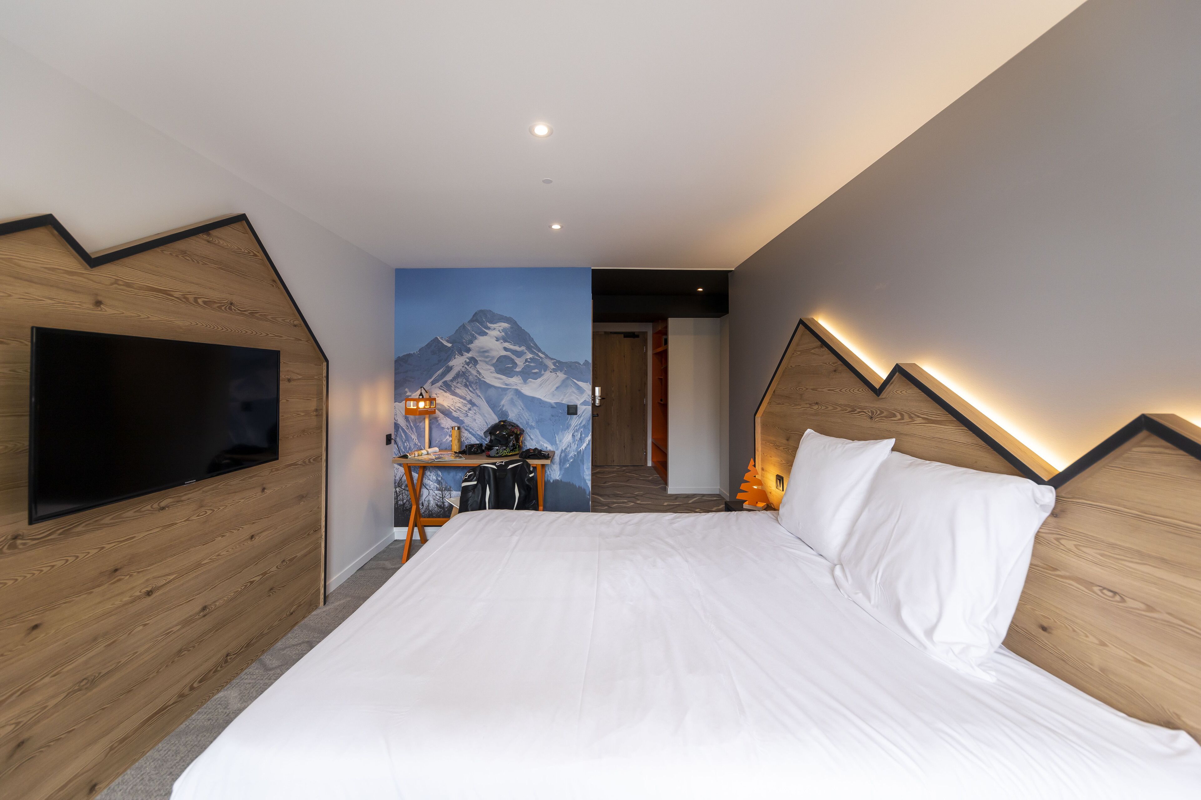 Une chambre d’hôtel moderne dotée d’un grand lit, d’un téléviseur à écran plat, d’un bureau avec une chaise et d’une fresque représentant une montagne sur le mur.